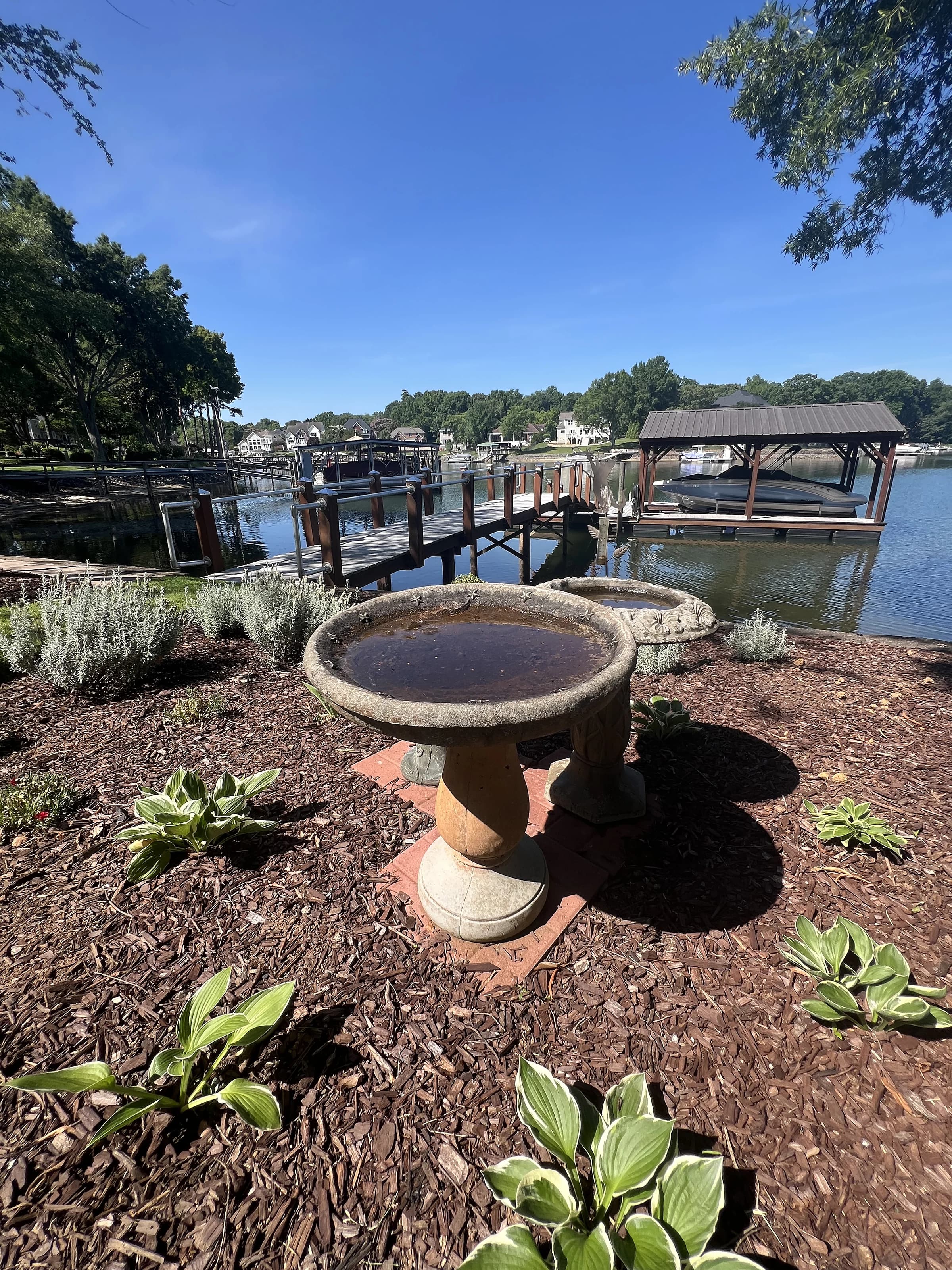 A stone birdbath set in a fresh mulch bed with hostas, overlooking a Lake Norman dock