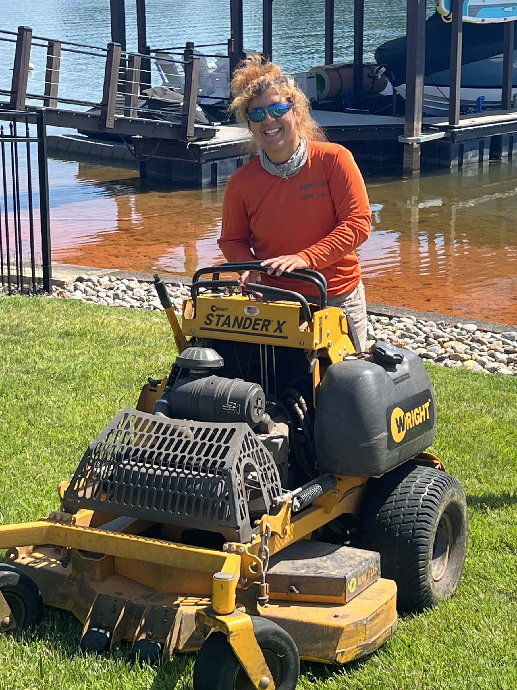 A Mow, Inc. crew member with a Wright stand-on mower on a lakefront lawn beside a boat dock